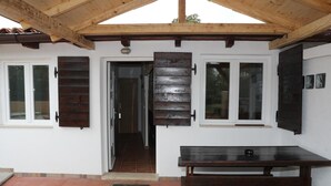 Ein überdachter Patio-Bereich mit einer Holzbank und einem Tisch, weißen Wänden und einer Tür mit Holzjalousien.