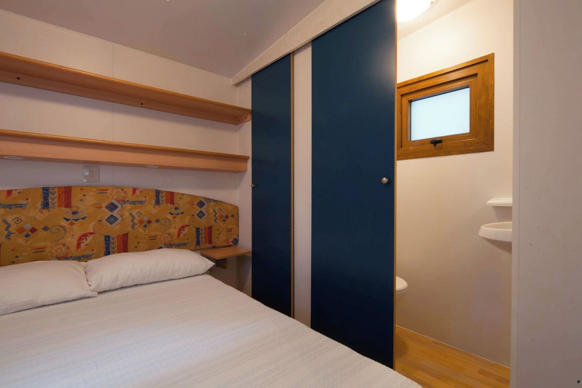Une chambre à coucher avec un lit, une tête de lit ornée d’un motif coloré, une étagère en bois, une porte bleue et une petite fenêtre.