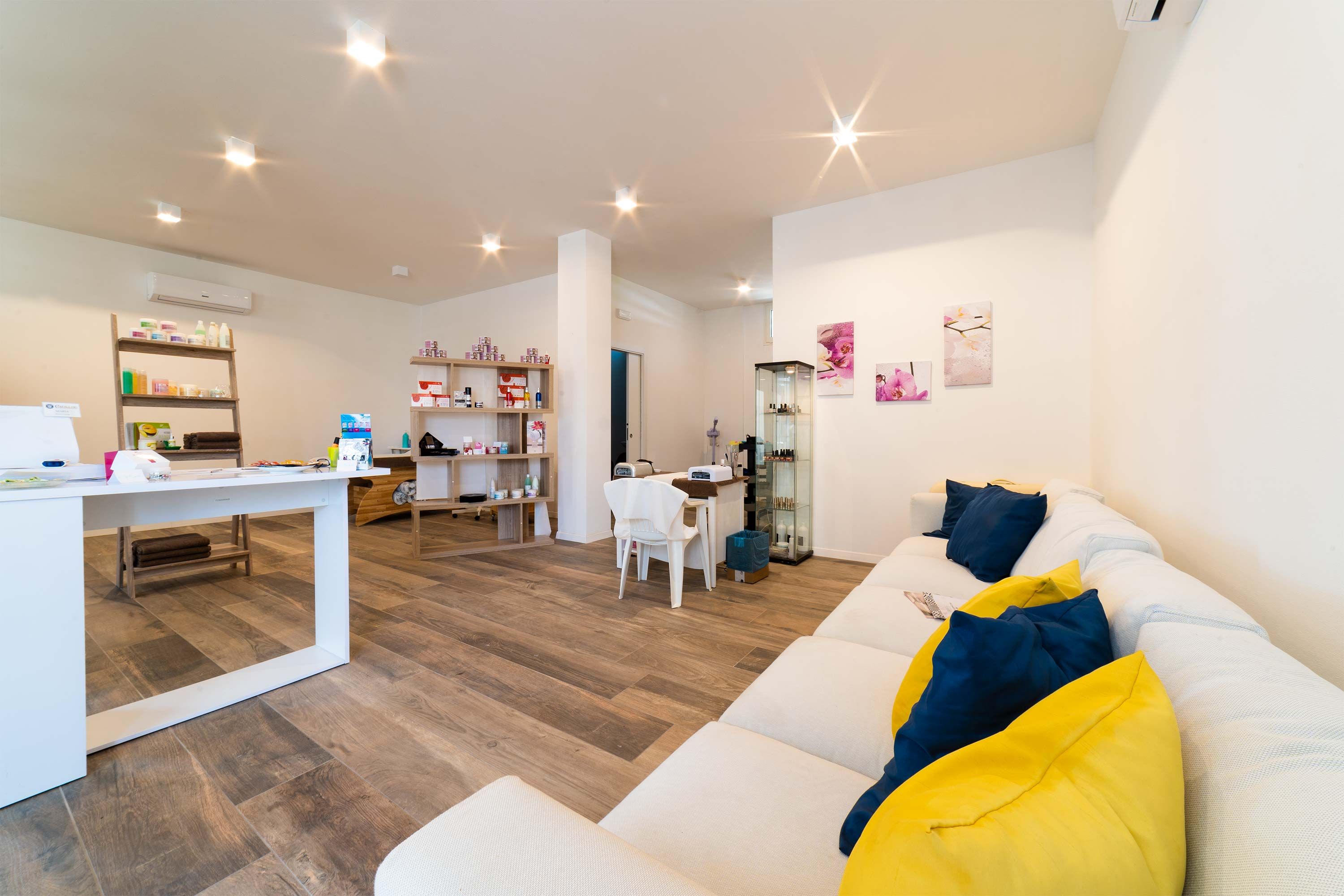 Un salon moderne avec un canapé blanc, des coussins jaunes et bleus, un coin repas avec une table et des chaises blanches, et un coin cuisine avec des étagères et des appareils électroménagers.