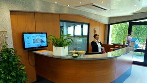 Una recepción con una pantalla de televisión, plantas en macetas y un ventanal con vistas a zonas verdes.