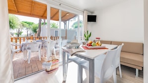A bright, open-plan living area with a wooden deck, white furniture, and a flat-screen TV mounted on the wall. - Camping Village Cavallino (Cavallino-Treporti)