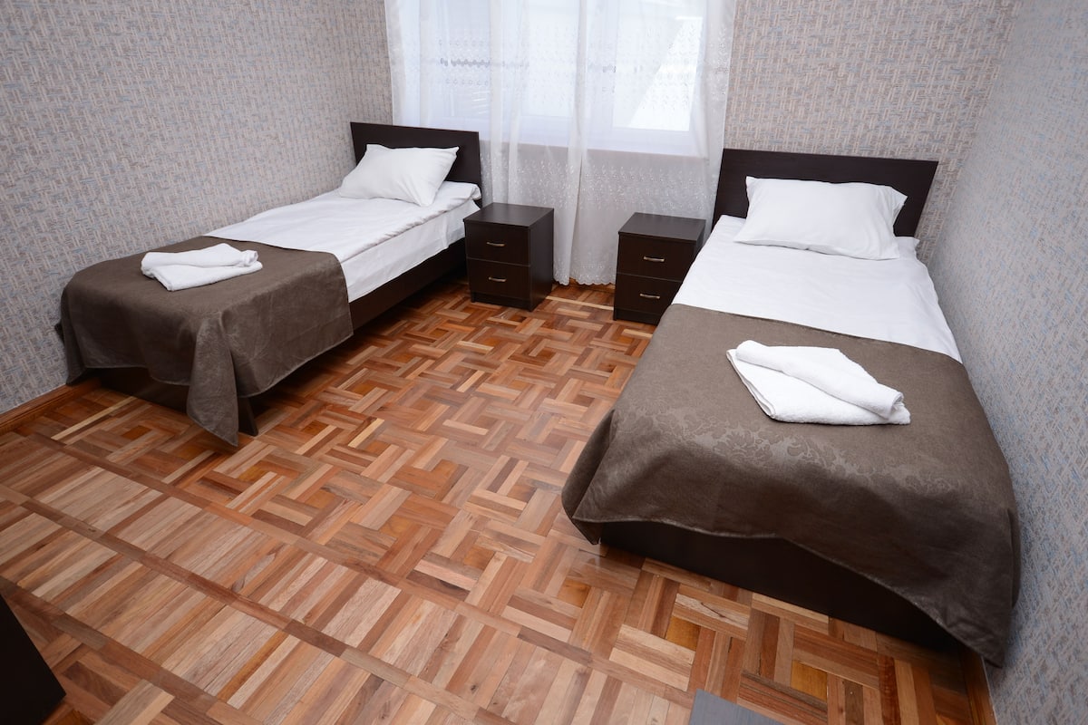 A hotel room with two beds, a herringbone-patterned wooden floor, and a window with sheer curtains.