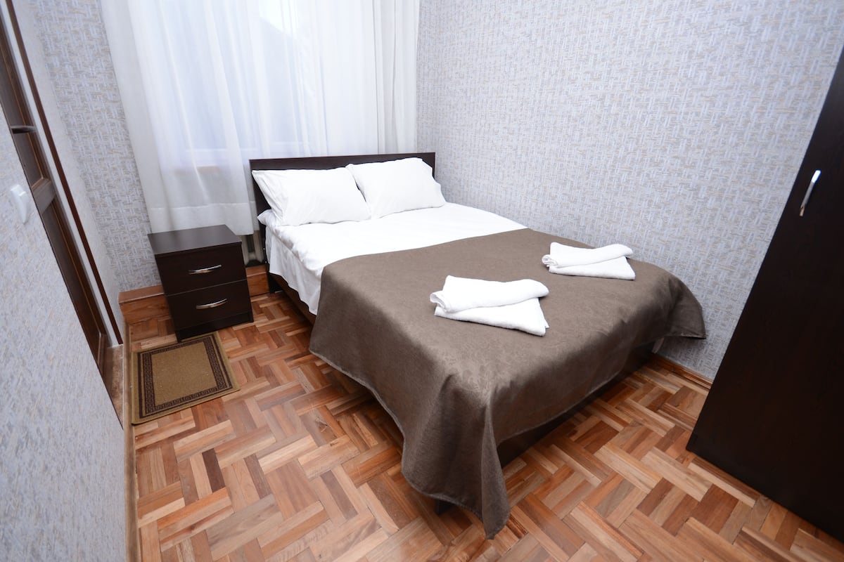 A neatly made bed with white linens and a brown bedspread, a wooden nightstand with a drawer, and a wooden herringbone floor.