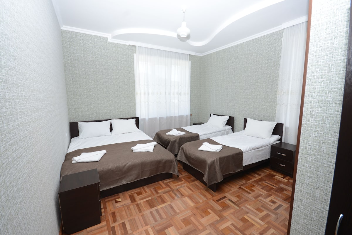 A hotel room with two beds, a wooden parquet floor, and a large window with sheer curtains.
