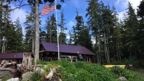 Un chalet doté d’un toit en métal, entouré d’une forêt dense et surmonté d’un mât portant le drapeau américain.