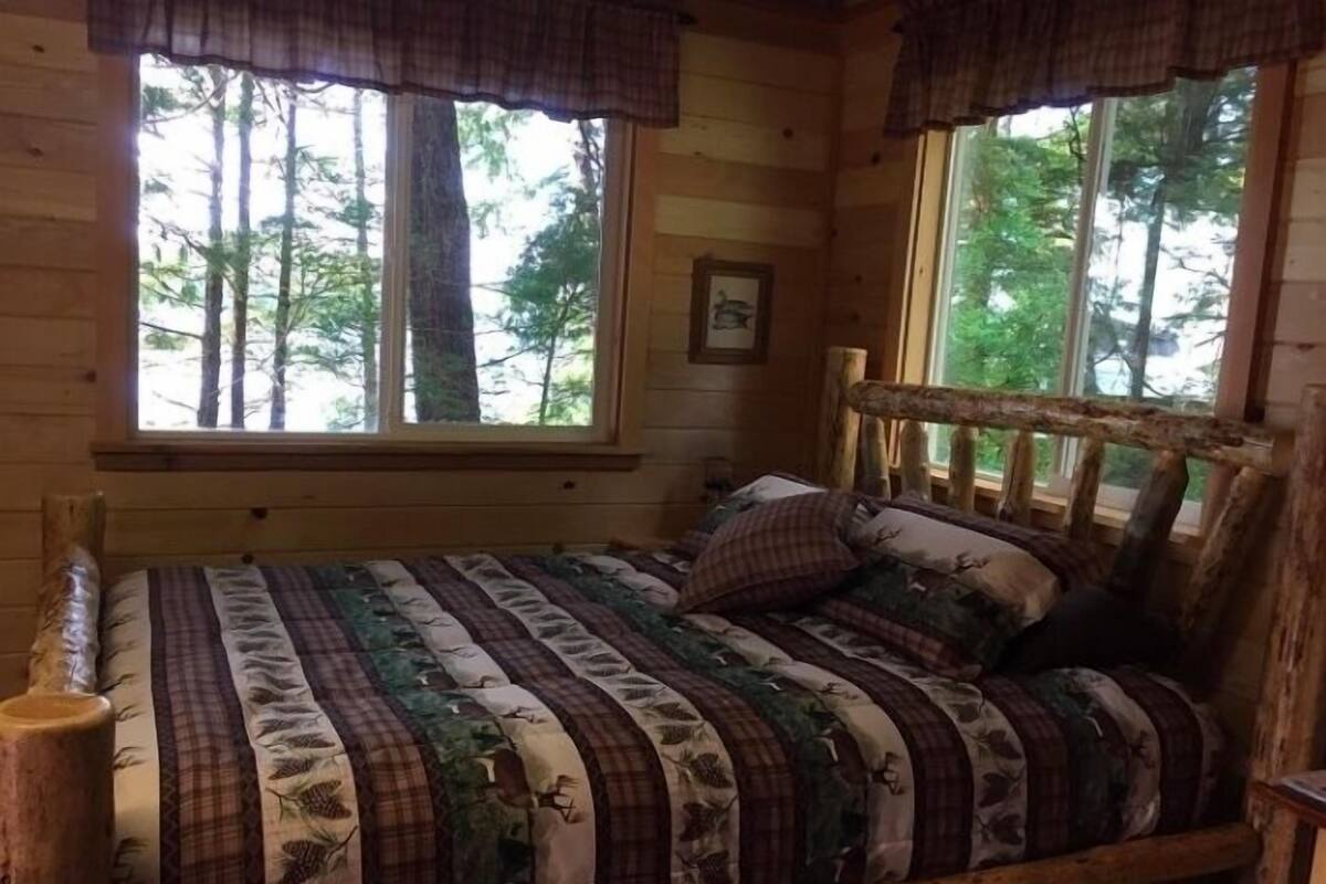 Ein gemütliches Blockhauszimmer mit einem hölzernen Bett, karierter Bettwäsche und großen Fenstern mit Blick auf den Wald.