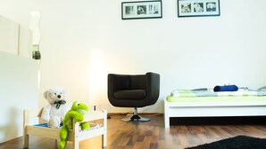 Una cama infantil con colchón blanco y manta verde, una silla negra, una mesita de madera con un osito de peluche y un juguete verde, y una alfombra negra en el suelo.