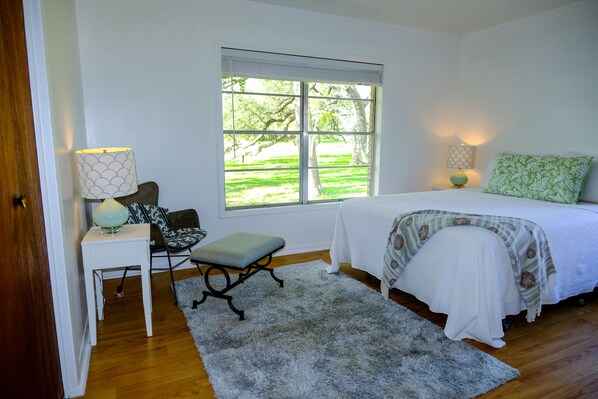 A bedroom with a bed, a window with a view of greenery, a small table with a lamp, and a chair.