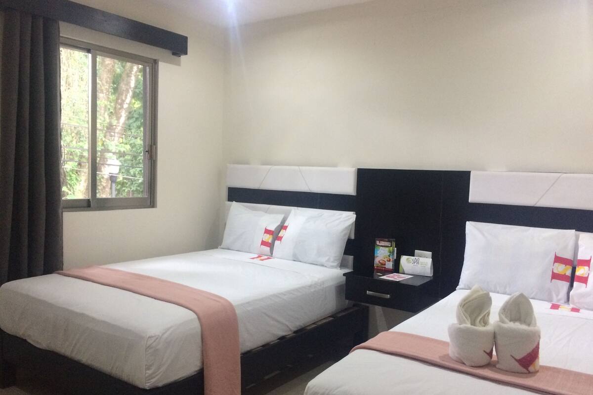 Habitación de hotel con dos camas, cada una con ropa blanca y una manta rosa. Se observa un mesita de noche con un libro y una botella de agua. Una ventana con cortinas es visible, y un par de pantuflas están sobre el suelo.