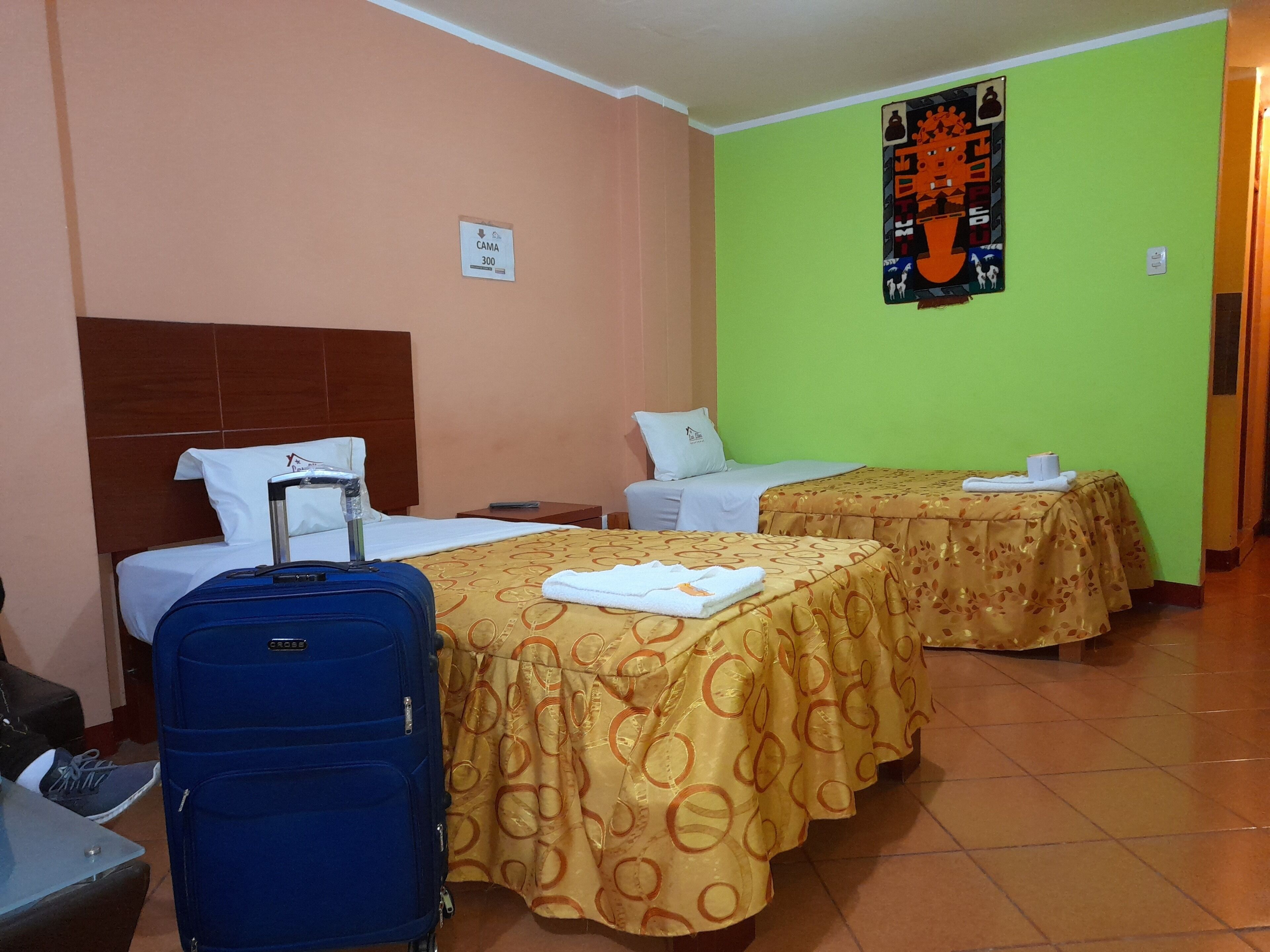 A hotel room with two beds, a blue suitcase, and a green wall with a poster.