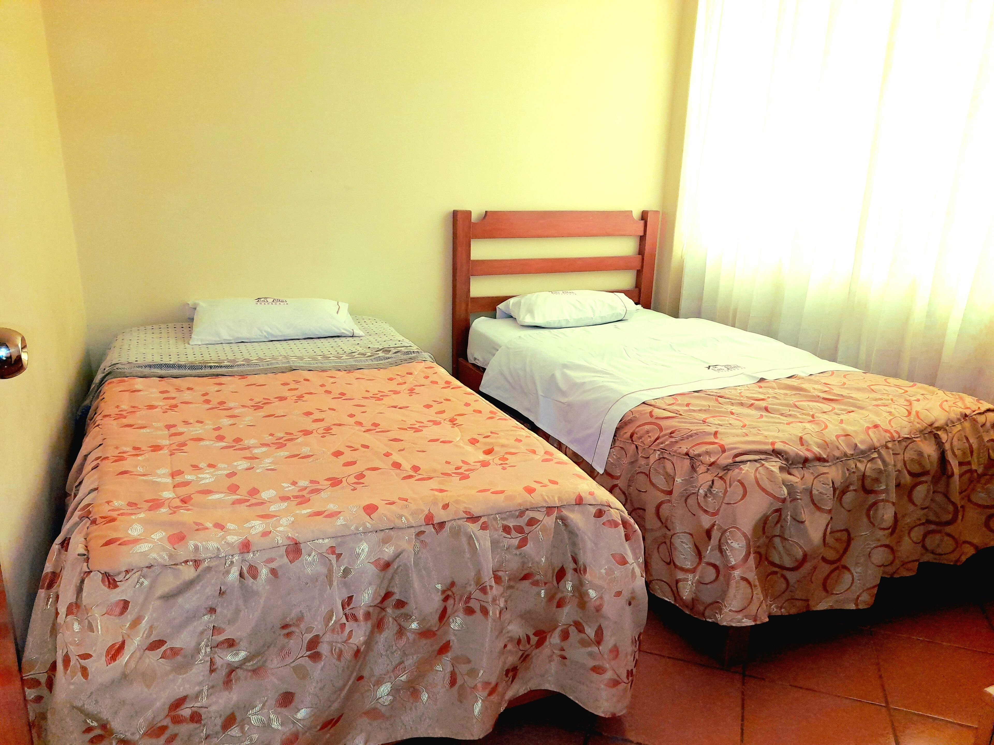 Two single beds with wooden headboards, a plain wall, and a window with curtains.