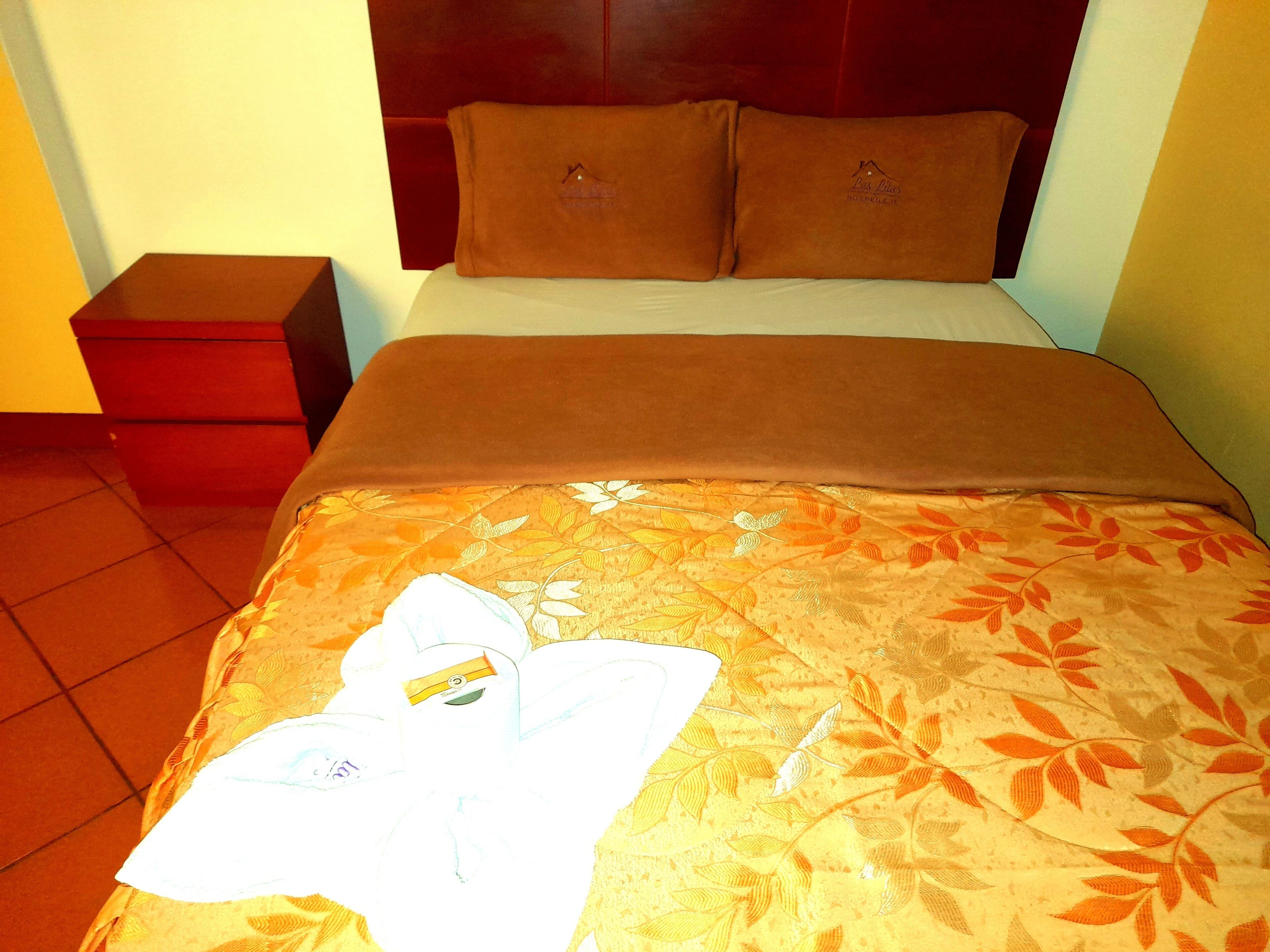 Una cama bien hecha con una colcha estampada con motivos florales, dos almohadas y una mesita de noche con un cajón rojo.