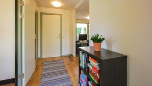 Een gang met een houten vloer, een gestreept tapijt, een boekenplank met boeken en een plant in pot, en een deur die naar een andere kamer leidt.