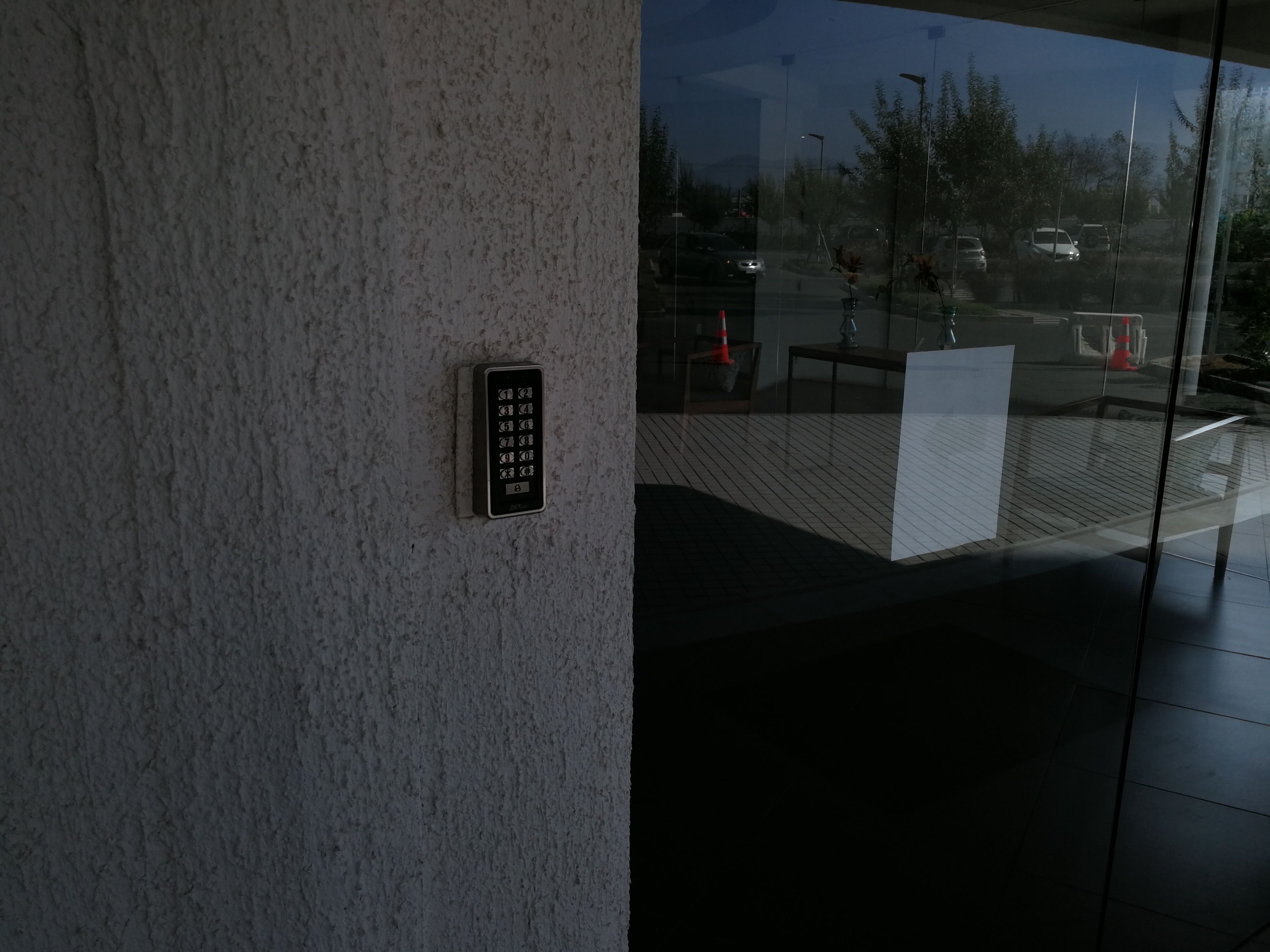 A keypad entry system on a textured wall, a glass door reflecting an outdoor area with a traffic cone and a vehicle.