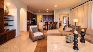 A spacious living area with a sofa, armchairs, and a coffee table. There is a kitchen in the background with a dining area. - Paraiso Del Mar Resort V58 4 Bed by Casago (La Paz)
