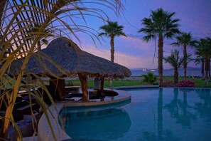 A pool area with a thatched-roof structure, palm trees, and a view of the sea.