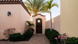 A courtyard with a tiled pathway leading to a dark wooden door, surrounded by lush greenery and a palm tree. - Paraiso Del Mar Resort V1 3 Bed by Casago (La Paz)