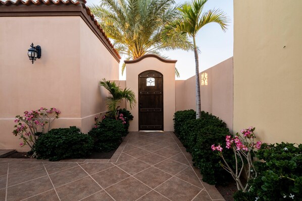 A courtyard with a tiled pathway leading to a dark wooden door, surrounded by lush greenery and a palm tree.