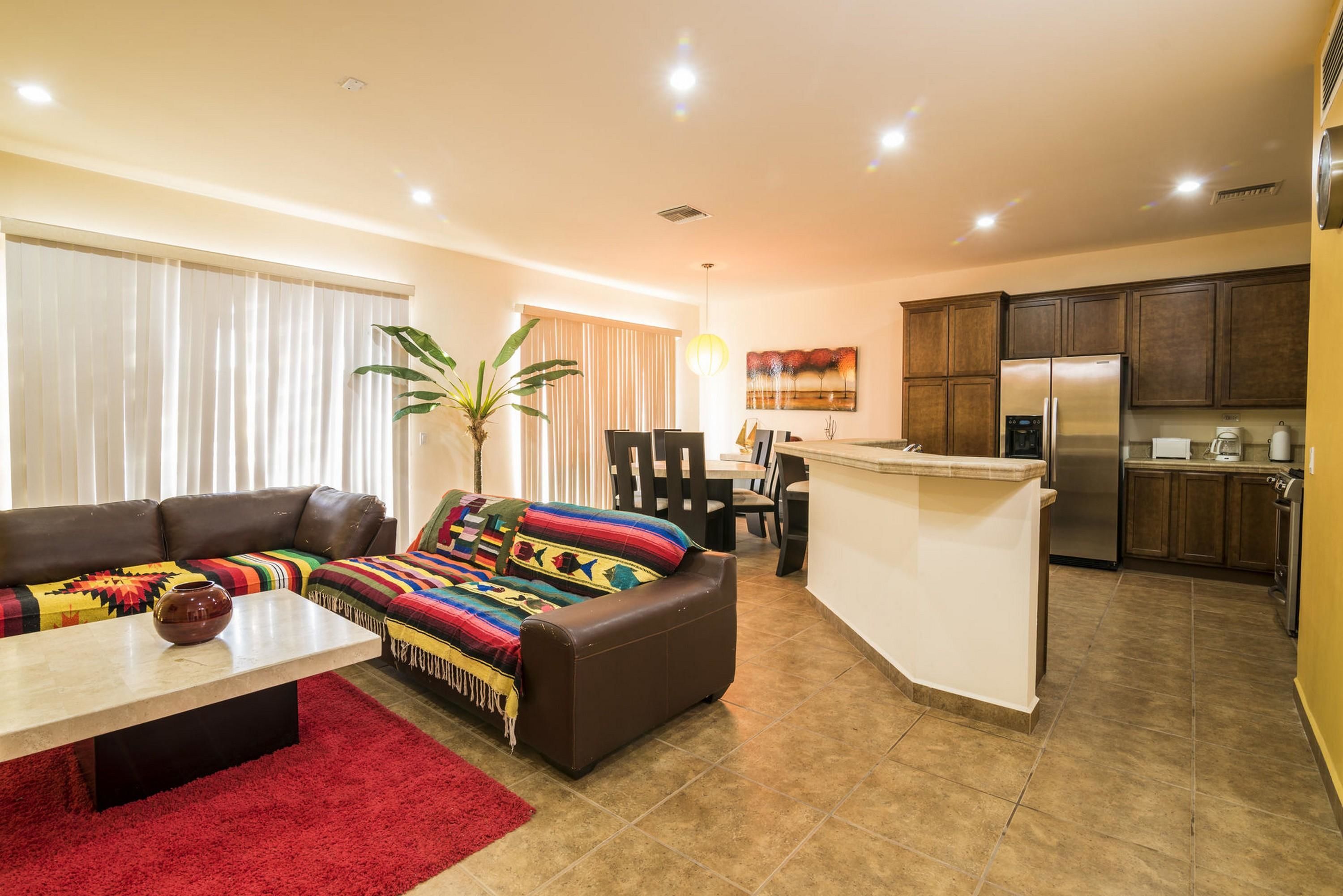 Una sala de estar moderna con un sofá, una mesa de centro y un tapete rojo. Hay una zona de cocina con armarios de madera y un refrigerador.