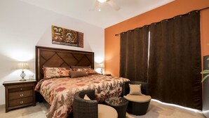 Un dormitorio con cama, mesitas de noche, sillas de mimbre, ventilador de techo y un amplio ventanal con cortinas.