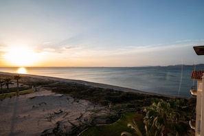 A beach view with a sunset, palm trees, and a building with a tiled roof. - Paraiso Del Mar Resort A502 3 Bed by Casago (La Paz)