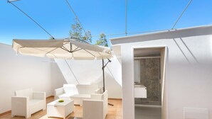Eine sonnendurchflutete Terrasse mit weißen Korbmöbeln, einem großen Sonnenschirm und einer Tür zu einem Badezimmer.