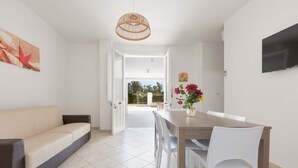 Ein modernes Wohnzimmer mit Sofa, Esstisch und Stühlen. Ein Fernseher ist an der Wand befestigt und ein großes Fenster bietet einen Blick ins Freie.