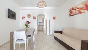 Ein modernes Wohnzimmer mit einer Couch, einem Esstisch mit Stühlen, einem an der Wand montierten Fernseher und einer Küche im Hintergrund.