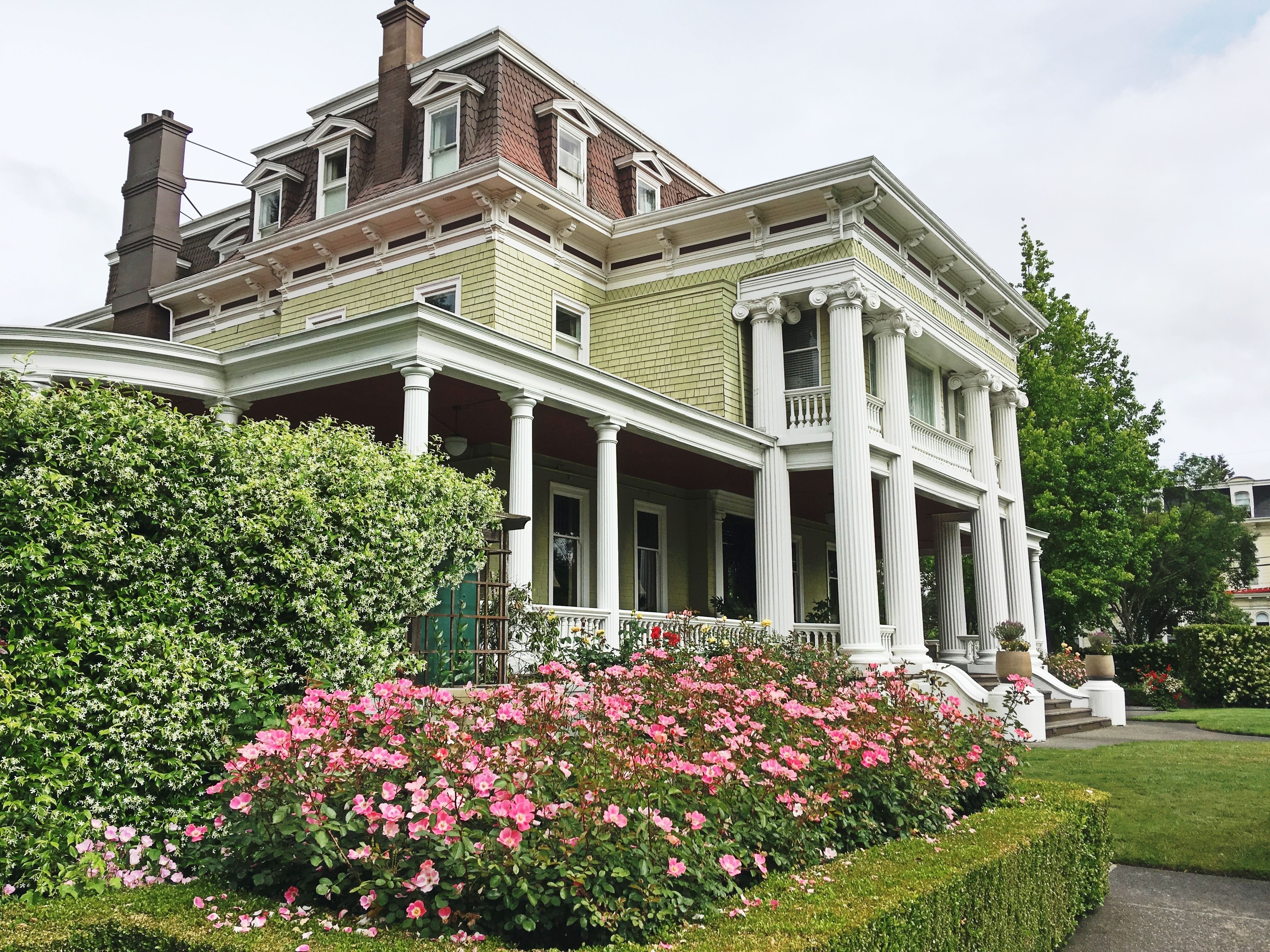 Una casa grande e histórica con un porche destacado, rodeada de abundante follaje y plantas florecientes.