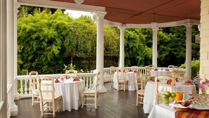 An outdoor patio with white columns and tables set for dining, surrounded by lush greenery. - Churchill Manor B&B (Napa)