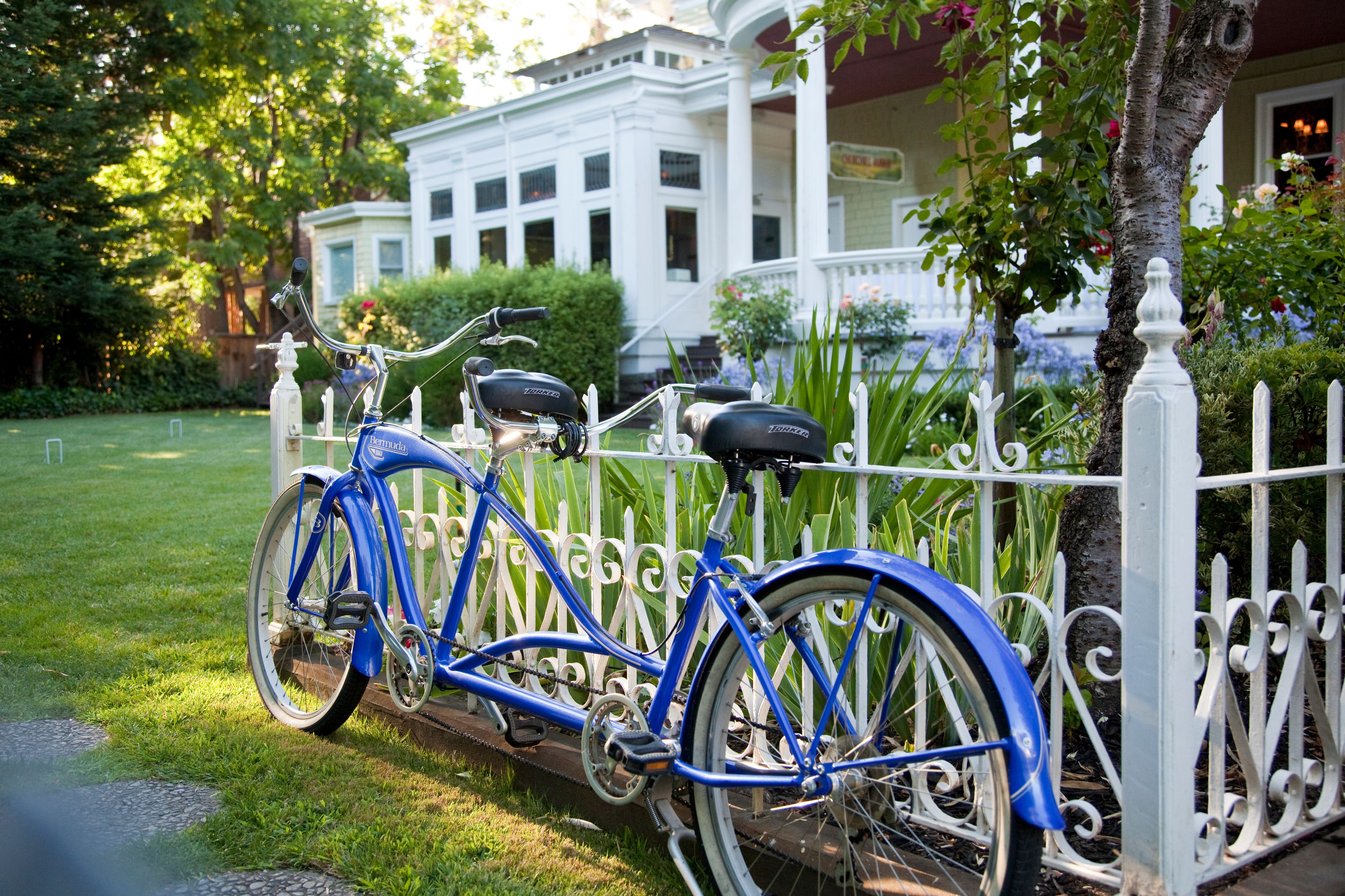 Uma bicicleta azul estacionada em frente a uma cerca branca, com uma casa ao fundo.