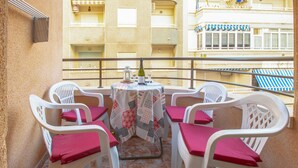 Un balcon avec une table dressée pour deux personnes, des chaises en plastique blanches avec des coussins rouges et une nappe à motifs.