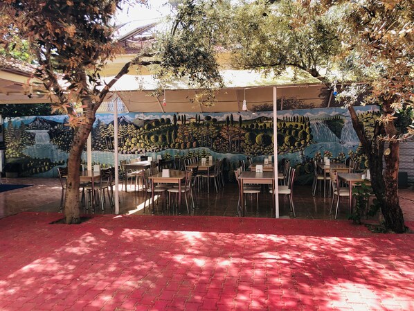 An outdoor dining area with tables and chairs, a mural on the wall, and trees providing shade.