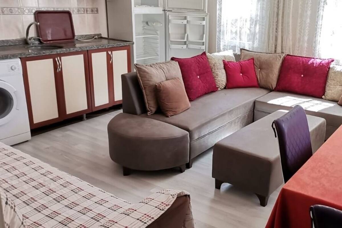A compact living space with a sofa set, ottomans, a dining table, and a kitchen area with cabinets and a washing machine.