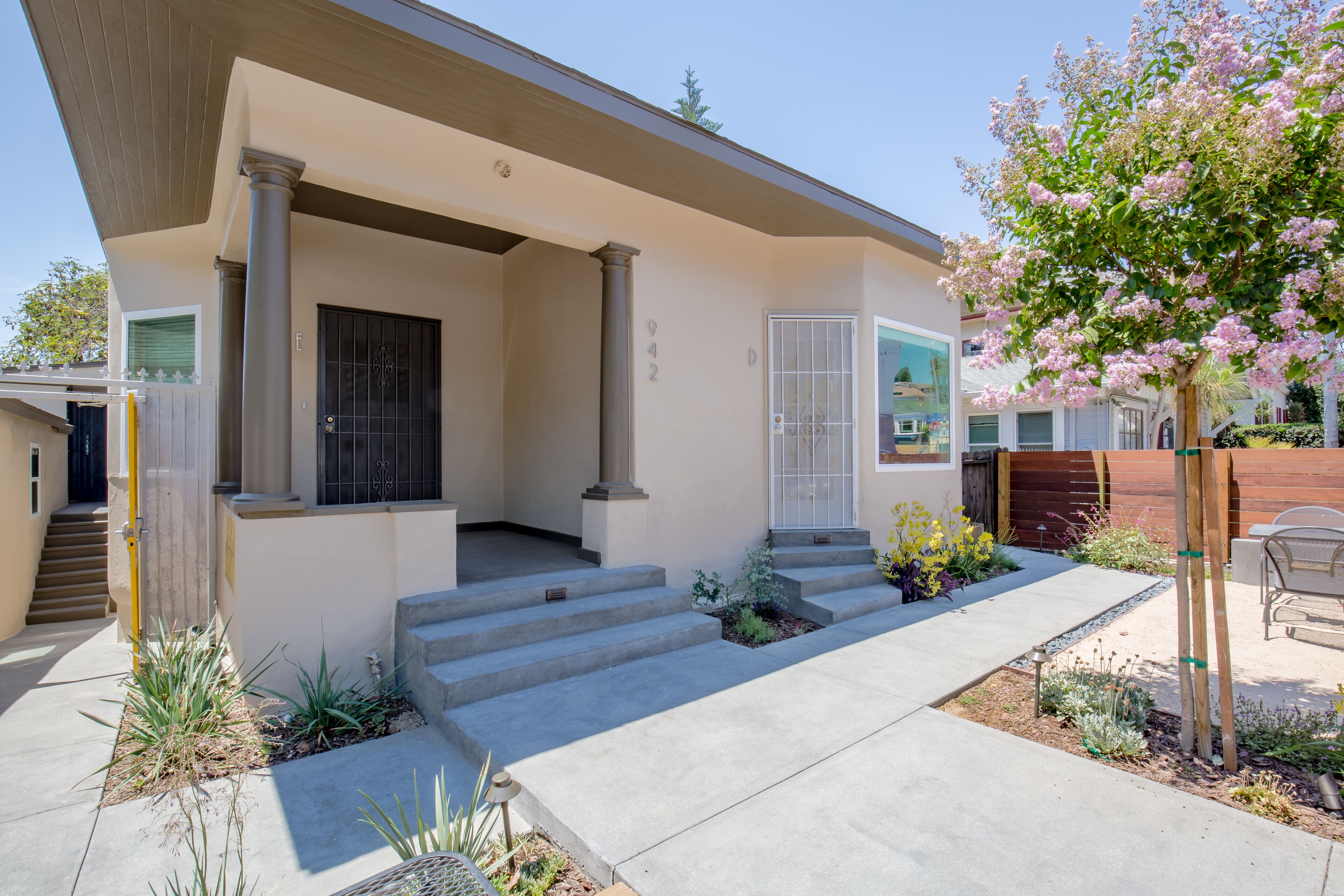 A modern house with a covered porch, a front door, and a small garden with plants and a tree.