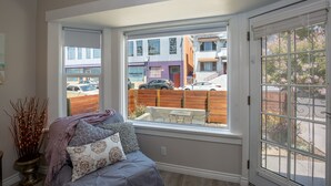 Ein Zimmer mit großem Fenster und Blick auf eine Straße, einem Bett mit Kissen und einem kleinen Tisch.