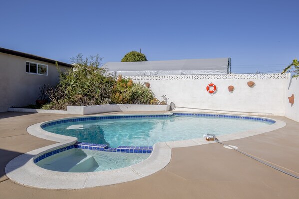 Una piscina con borde curvo, rodeada por una terraza de hormigón. Hay plantas y una pared blanca con detalles decorativos.