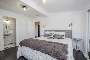 Un dormitorio moderno con una cama grande, una mesita de noche, un espejo y un baño visible a través de una puerta abierta.