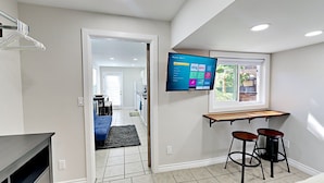 A modern kitchen with a built-in TV displaying streaming services, a window with a view, and a small bar area with stools. - Cool New Remodel Near Coronado, NEW Jacuzzi, Fire Pit, BBQ, Walk to Downtown! (San Diego)