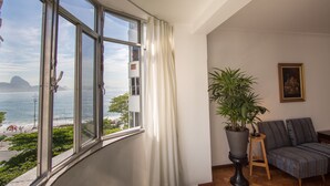 A room with a large window overlooking a beach, a potted plant, a sofa, and a framed picture on the wall. - LineRio Vista Mar Copacabana 228 (Rio de Janeiro)