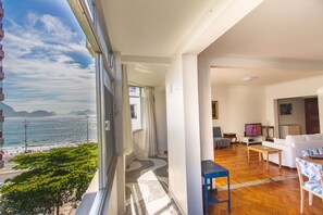 Ein Balkon mit Blick auf Meer und Berge, ein Wohnzimmer mit Sofa und Fernseher sowie ein Essbereich mit Tisch und Stühlen.