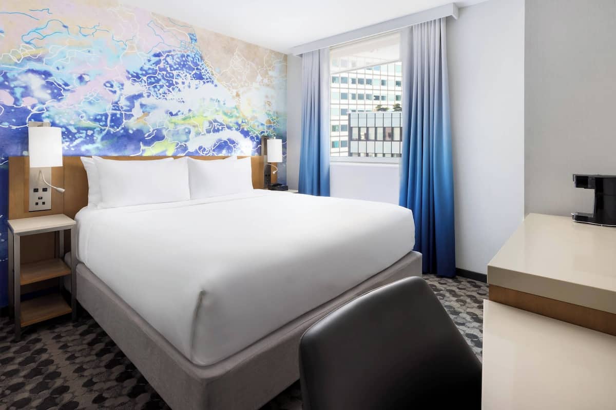 A hotel room with a large bed, a desk, a chair, a nightstand with a lamp, and a view of the city through the window.