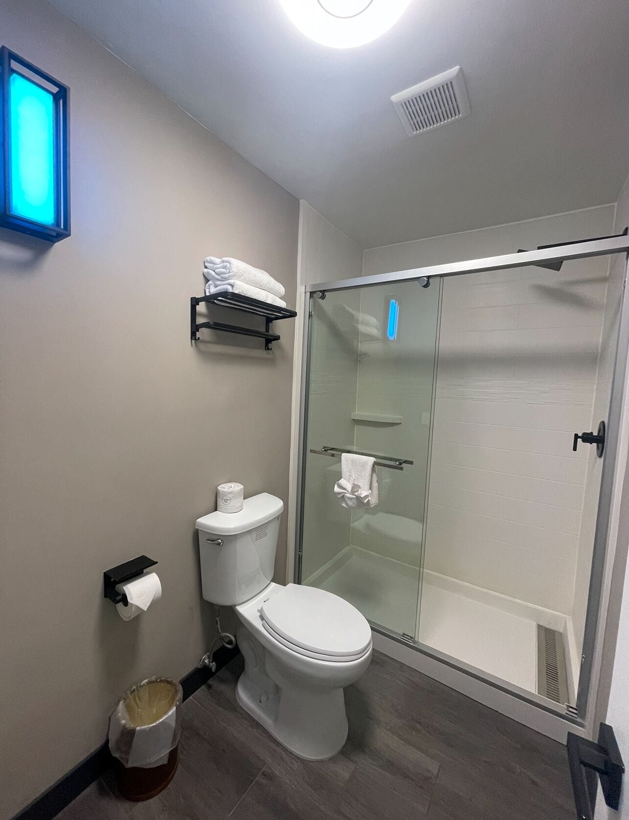 A modern bathroom with a toilet, a glass shower enclosure, a towel rack, and a wall-mounted shelf with towels.