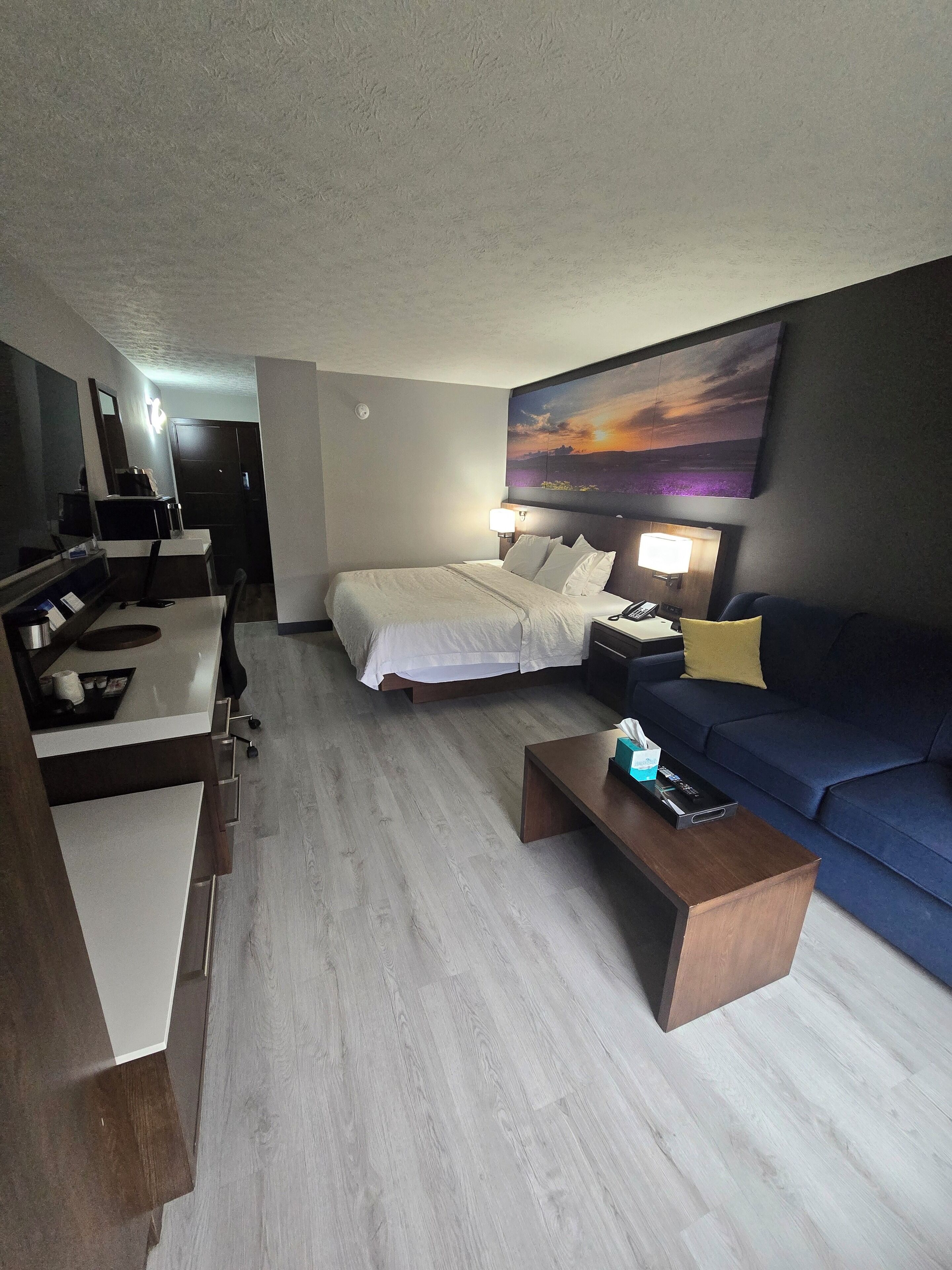 A modern hotel room with a bed, a sofa, a coffee table, and a desk with a lamp.
