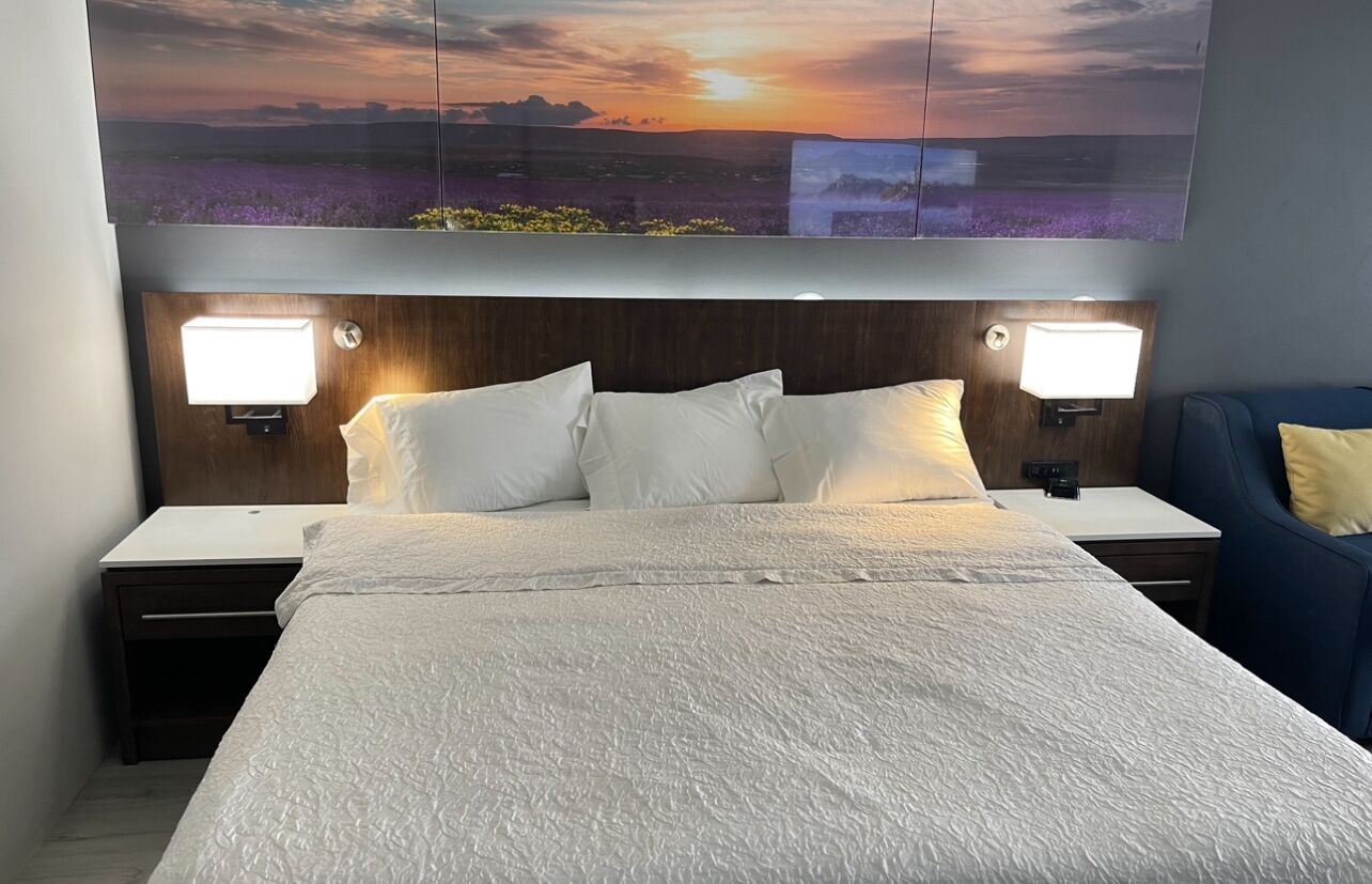 Een hotelkamer met een groot bed, twee nachtkastjes, een fauteuil en een aan de muur bevestigde televisie waarop een zonsondergang te zien is.