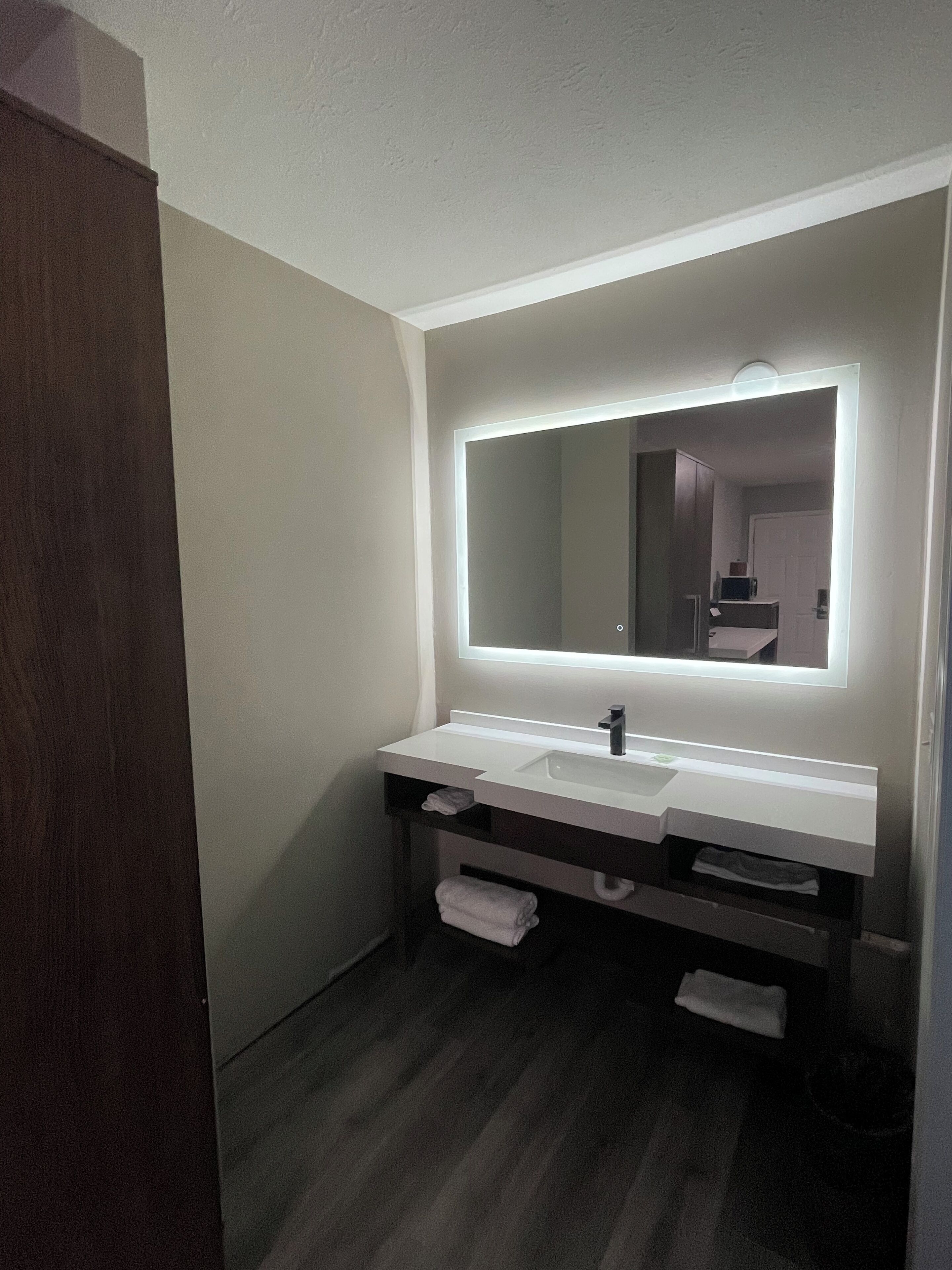 Un baño moderno con un tocador blanco, un espejo grande con iluminación integrada y un suelo de madera oscura.