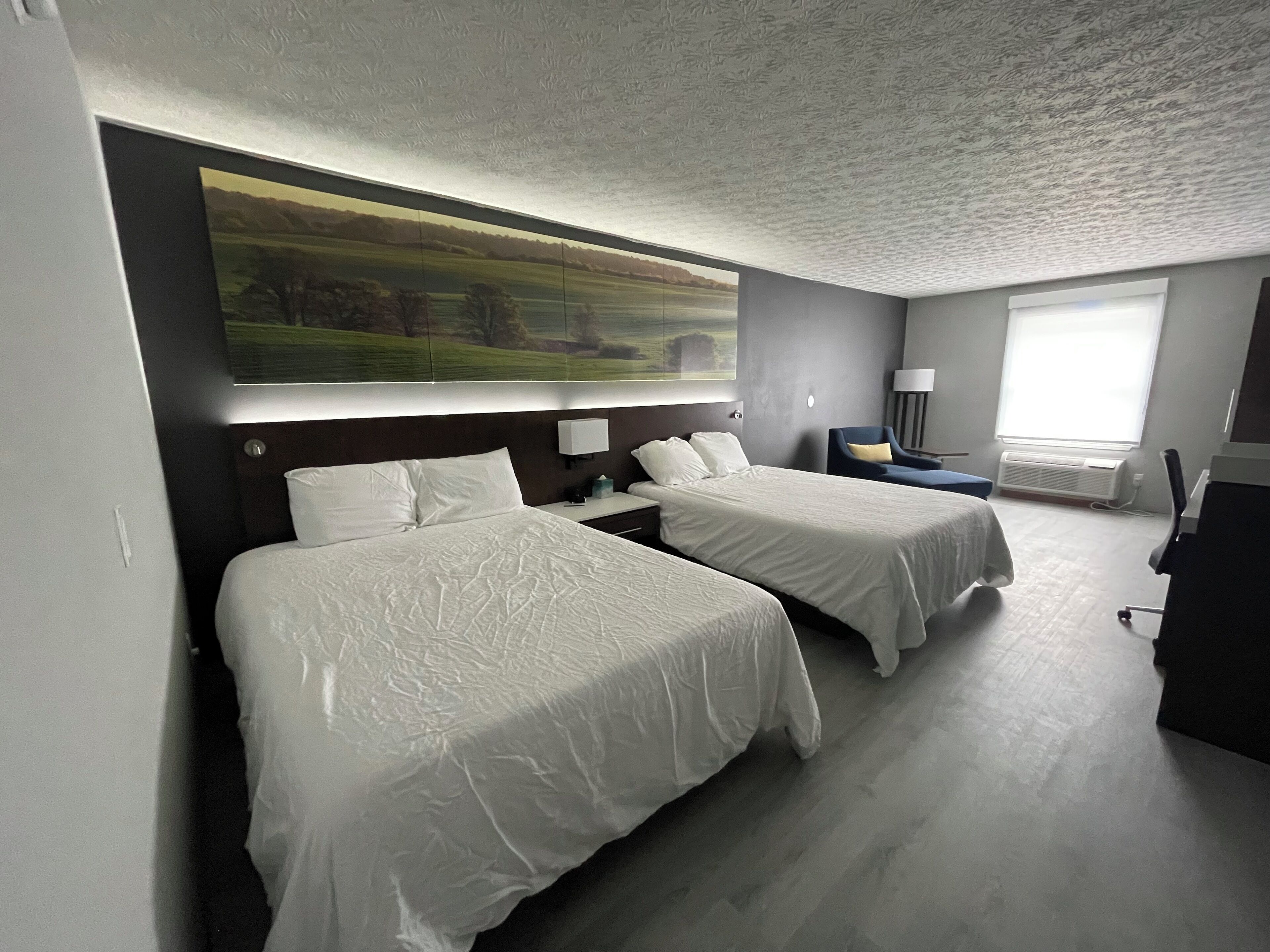 Een hotelkamer met twee bedden, een groot schilderij aan de muur, een bureau met een lamp, en een raam met airconditioning.