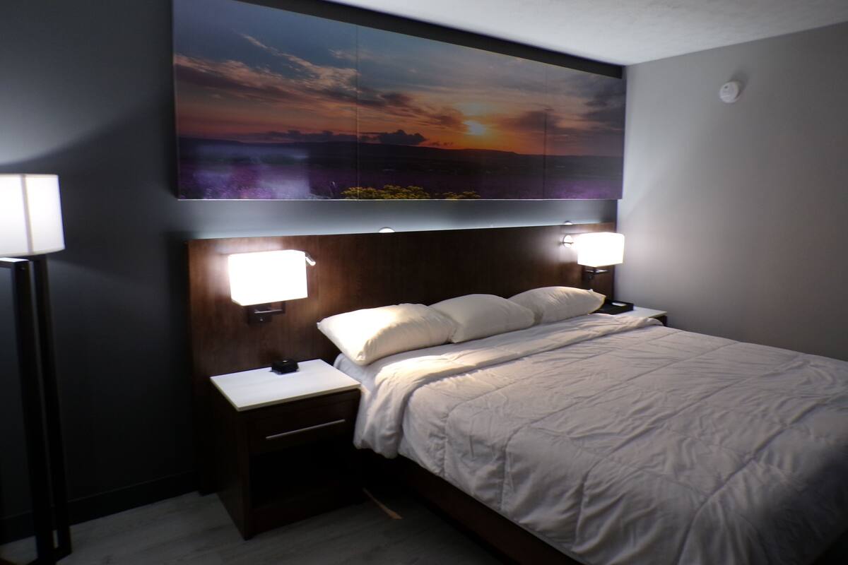 A hotel room with a large bed, two bedside lamps, a nightstand, and a wall-mounted TV displaying a sunset scene.