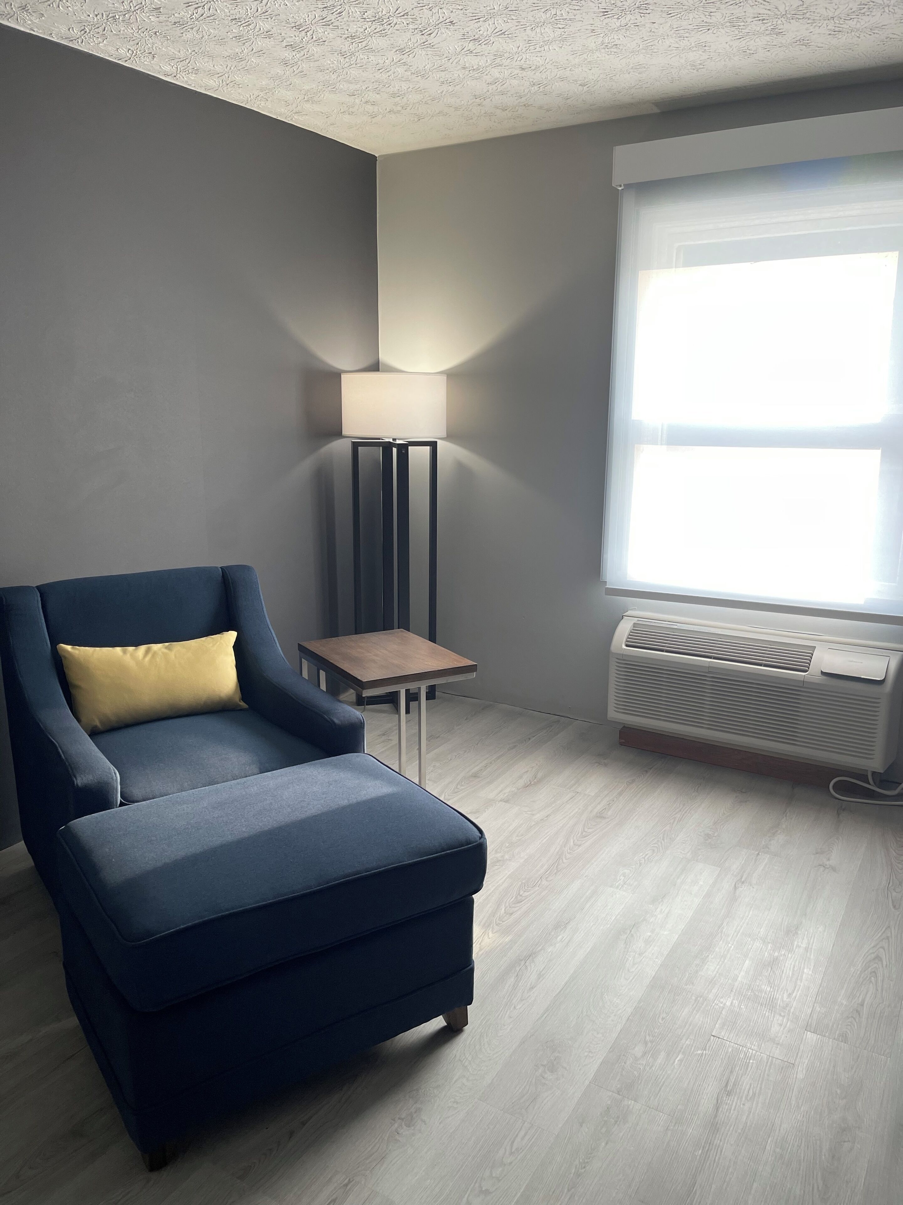 Een kamer met een blauwe fauteuil, een klein houten tafeltje, een vloerlamp en een raam met een airconditioner.