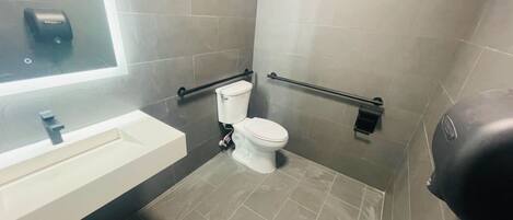 Un baño moderno con lavamanos blanco, inodoro y bidé. Hay barras de apoyo en las paredes y un dispensador de jabón sobre el lavamanos.